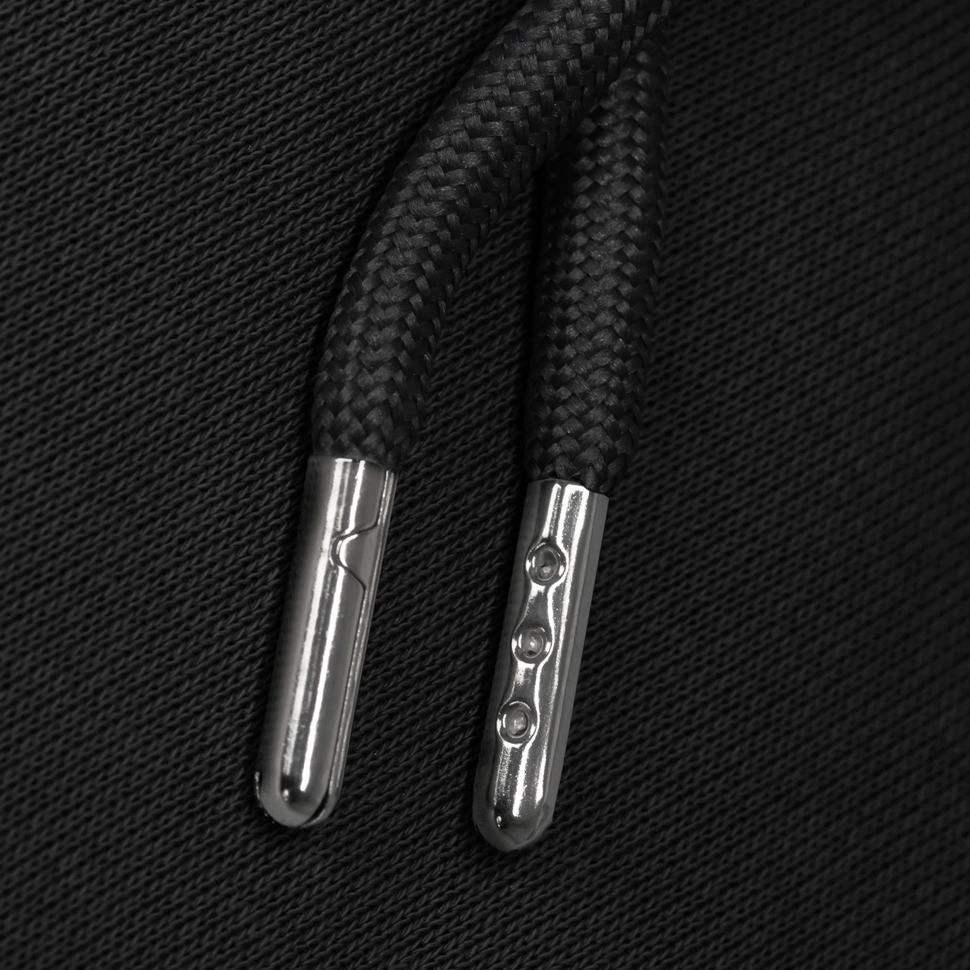 Black hoodie strings with silver aglets against a black fabric background.