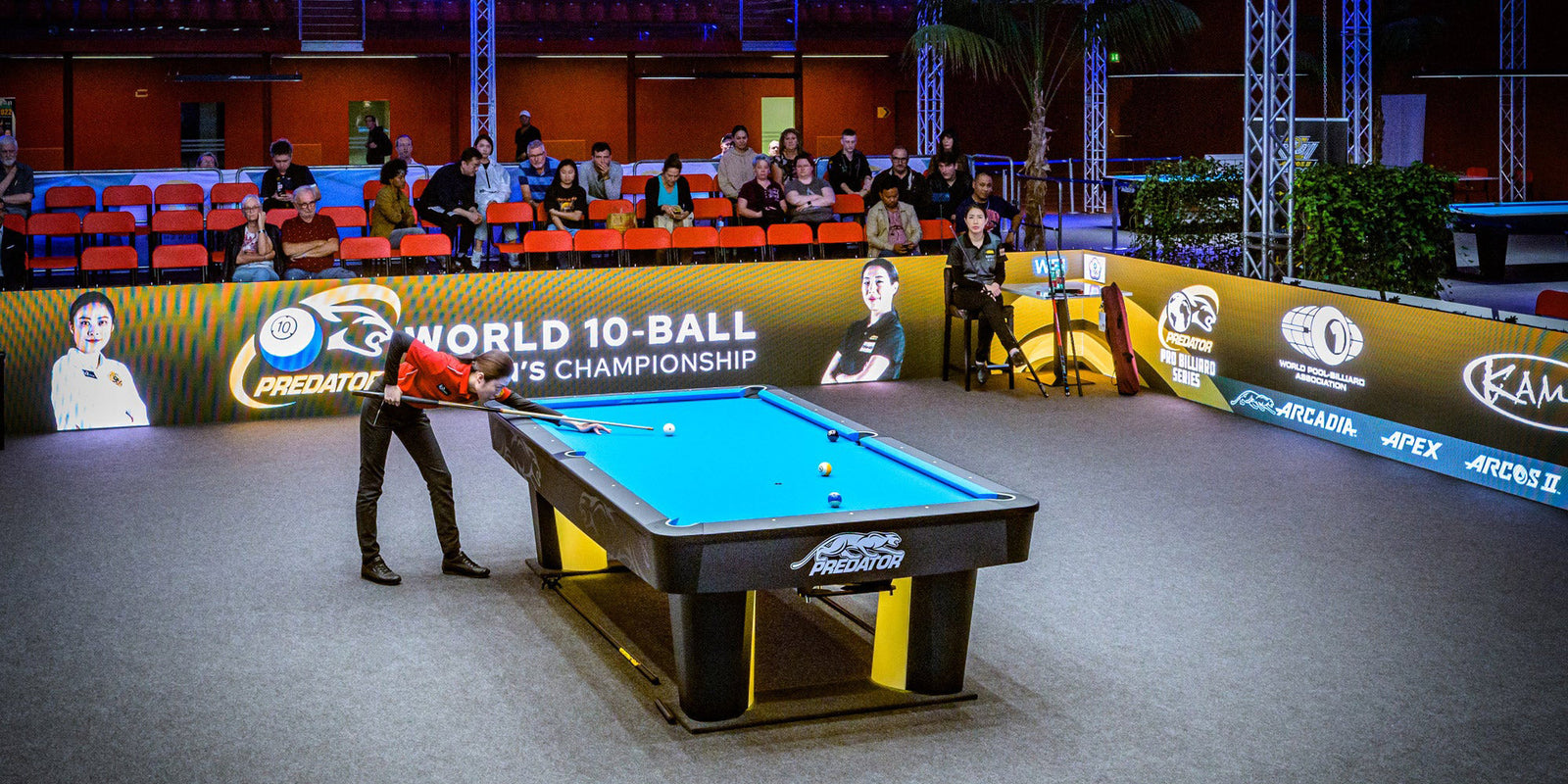 Pool table with a player in a red shirt, surrounded by spectators and event branding.