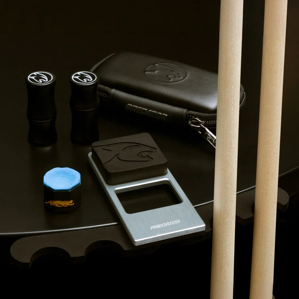 pool cue holder set up against a black table, with 2 cues resting against it. Its black carry case, a piece of chalk and joint protectors are set on the table next to it.