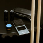 pool cue holder set up against a black table, with 2 cues resting against it. Its black carry case, a piece of chalk and joint protectors are set on the table next to it.